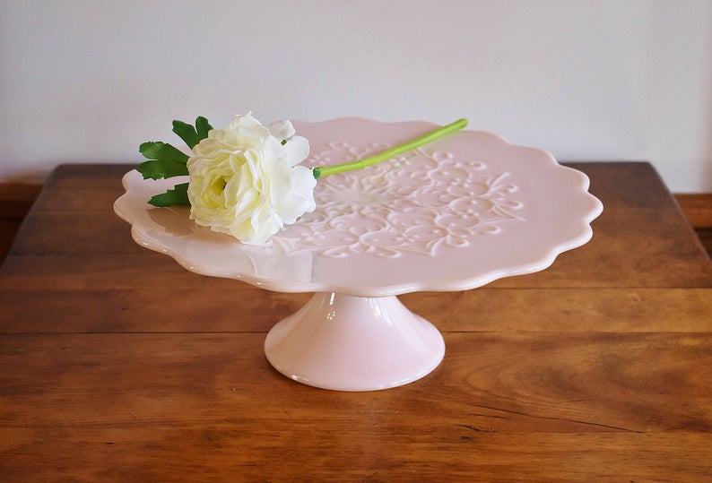 Blush pink milk glass cake stand standing on a wooden table with a white flower placed on top. 