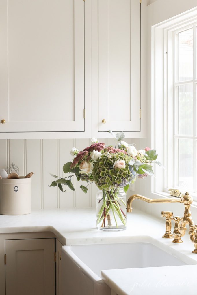 Beabdoard backsplash in an all white kitchen with brass accents and fresh flowers on the counter. 