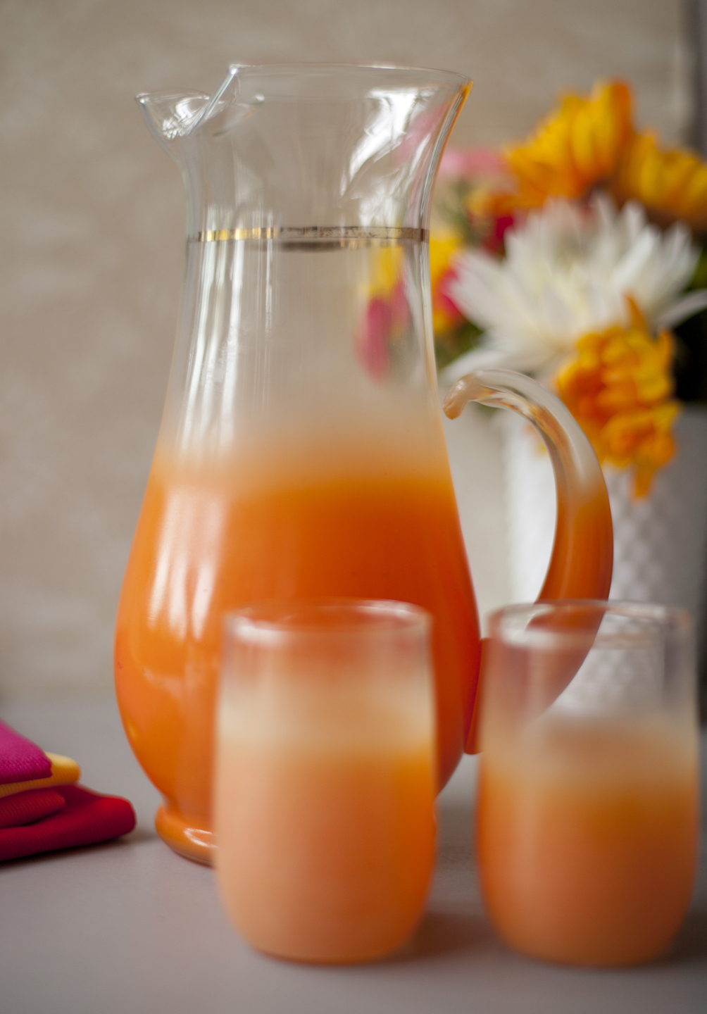 Bright orange Blendo Glassware pitcher and 2 tumblers placed near a vase of flowers. 