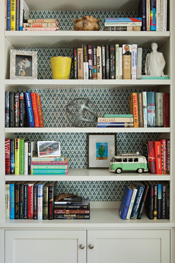 Bookshelf with patterned wallpaper in the background and filled with books and found objects. 