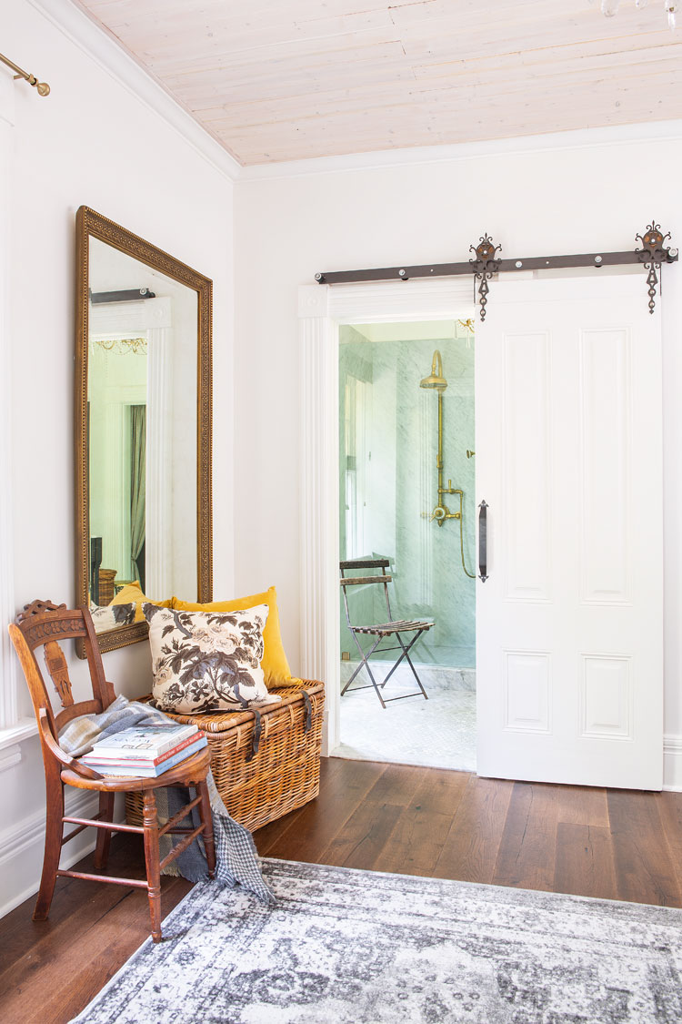 A lovely hallway with a small vignette of a whicker basket, large wooden mirror and antique wooden chair. The barn door is slid open you can see into the bathroom. 