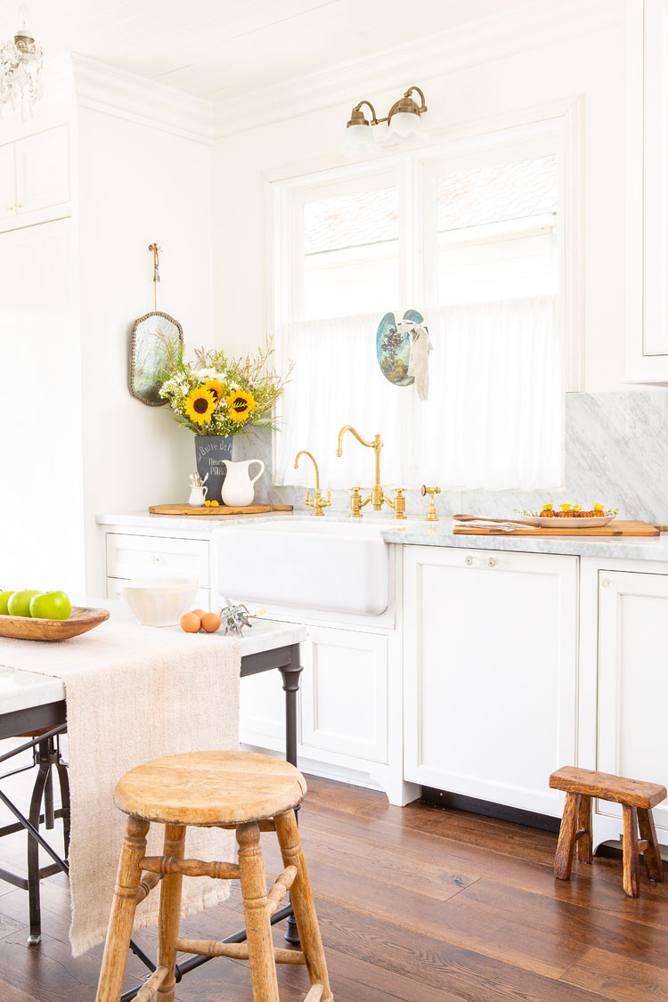 Bright and white kitchen sink with golden hardware at the farmhouse sink and wooden stools accenting the table and sink area. 