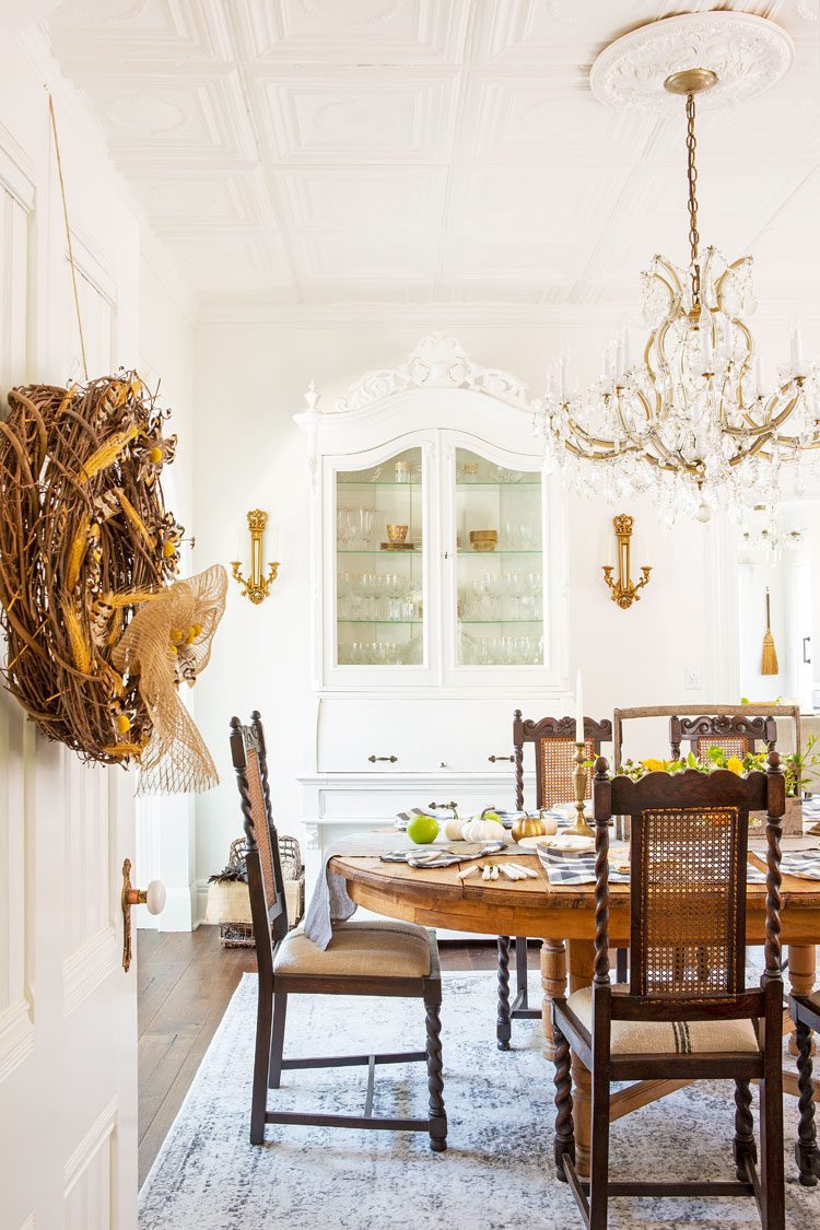 A peek in to the dining room, a wreath hanging on the door that you're peering through, a wooden, antique dining set fully ready to see meal service and a painted white china cabinet against the back wall. 