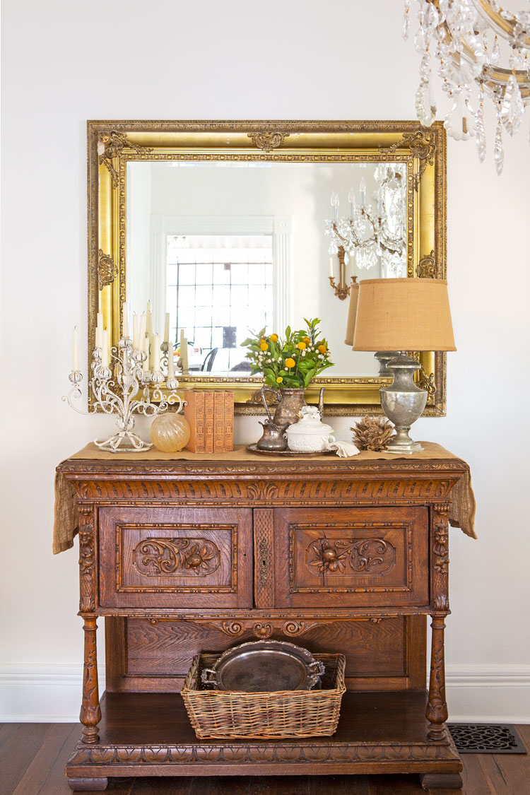 An ornate wooden hutch below a gold framed mirror. A crystal chandelier hanging from above and treasures displayed along the top of the hutch. 