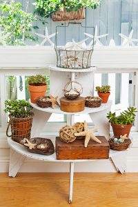 a white wooden plant stand with 3 tiered shelves each one larger than the other from top down. The shelves are filled with plants and decorative boxes