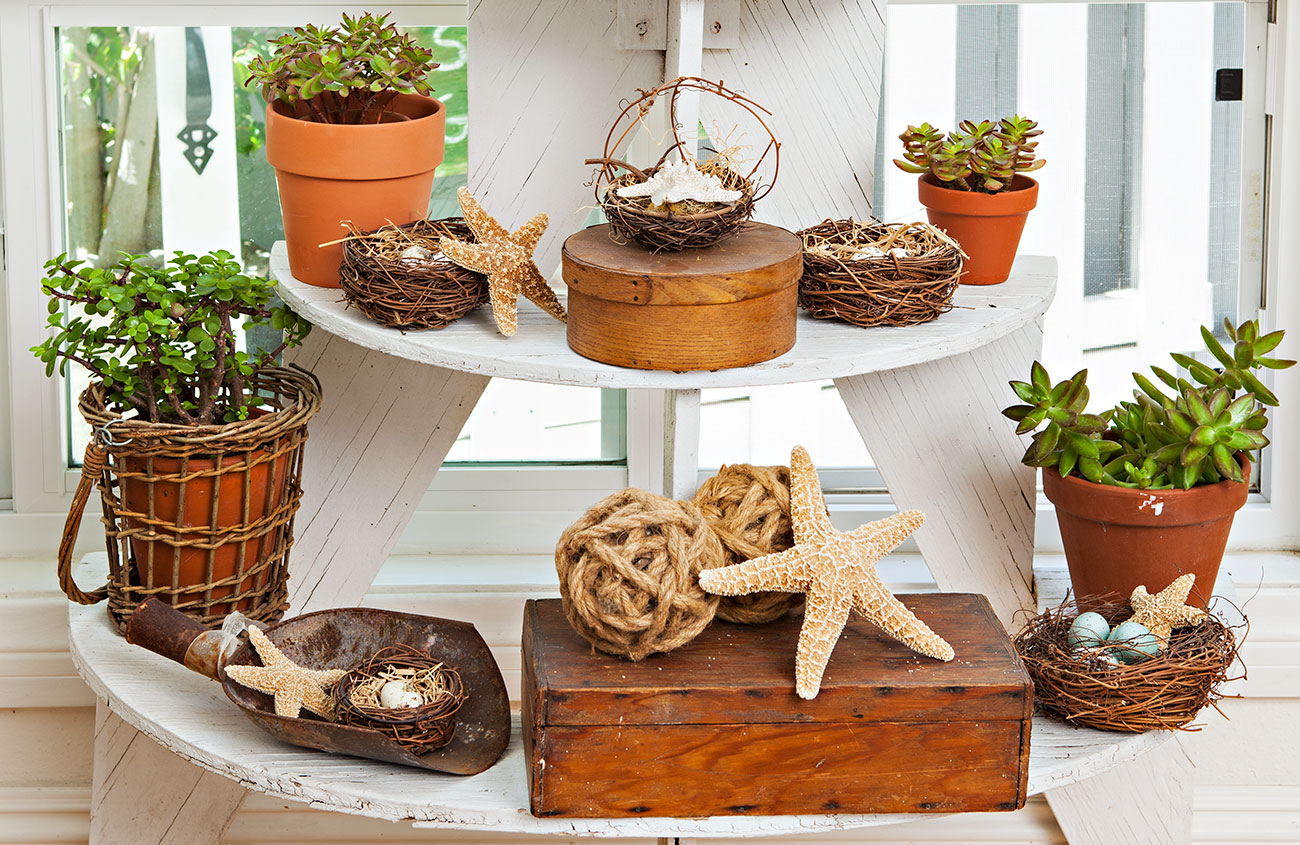 a white wooden plant stand with 3 tiered shelves each one larger than the other from top down. The shelves are filled with plants and decorative boxes