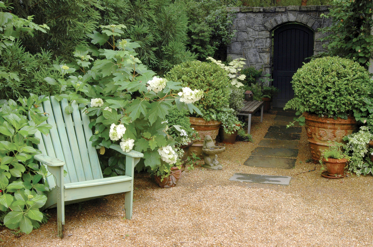 A light green Adirondack chair surrounded by an overgrown garden near a path leading to a stone wall with a black door.