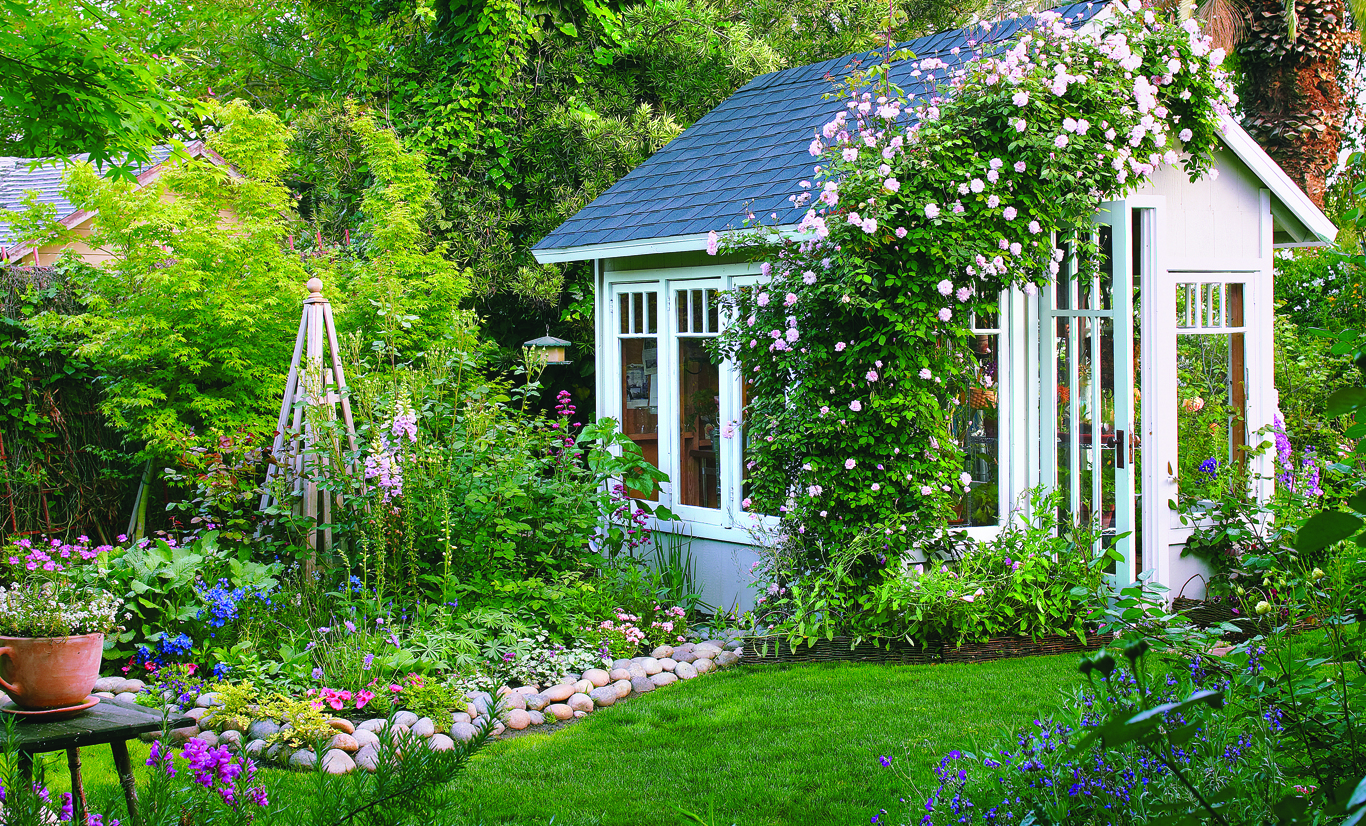 Vibrant green yard with a cottage style shed. The open shed door is welcoming and beautiful flowers grow up the side of the wall tying the shed into the yard.