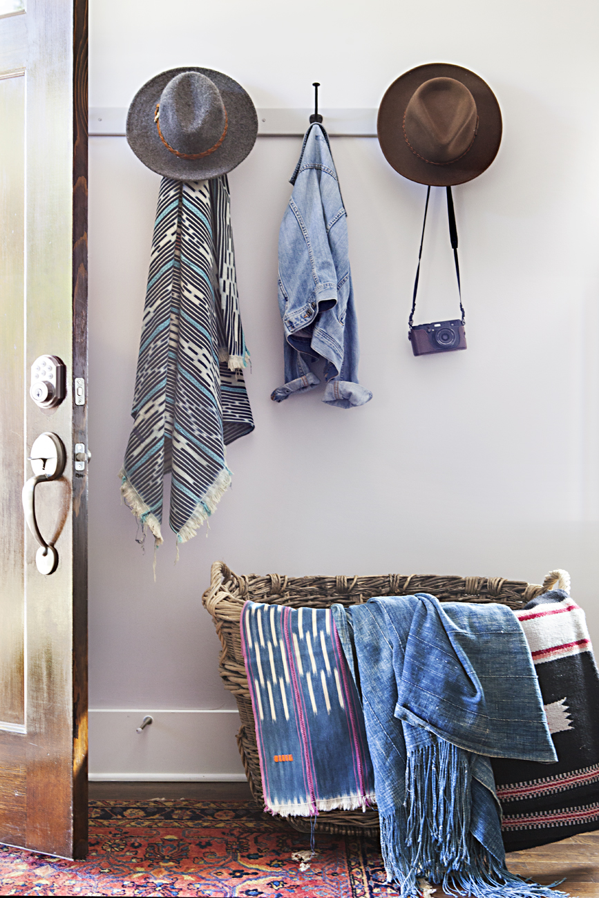 Textiles hanging over a woven basket and hats hanging on row of hooks behind the front door. 