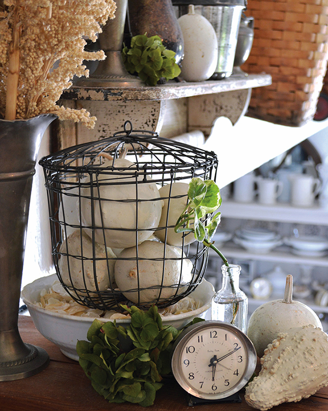 Wire cage filled with dried, cream colored pumpkins placed around a cottage as decor next to a shelf and an urn of dried flowers. 