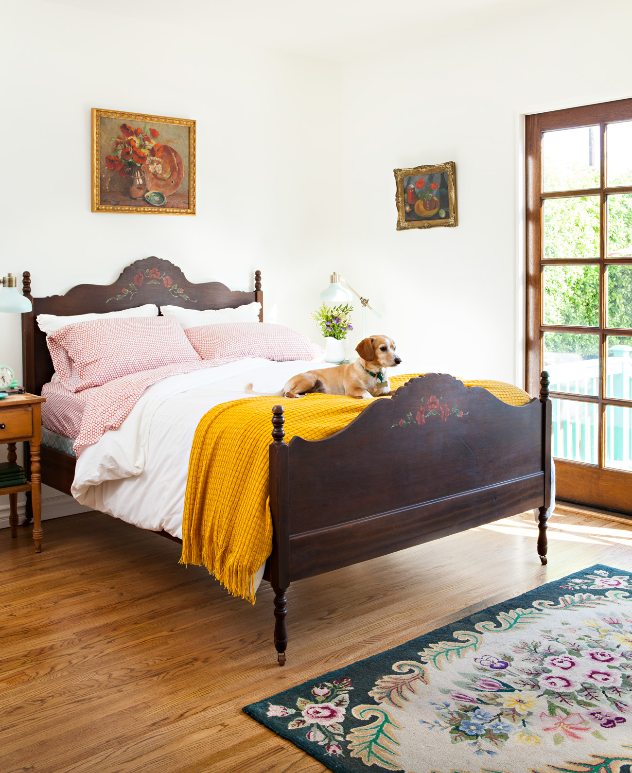 Antique bed in a spanish bungalow with vintage art on the walls and a yellow throw blanket on the end of the bed with a puppy on top. 