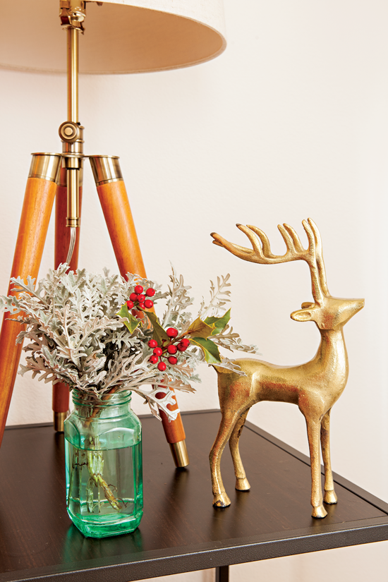 A green mason jar filled with seasonal shrubbery is placed alongside a small gold deer figure situated on a side table.