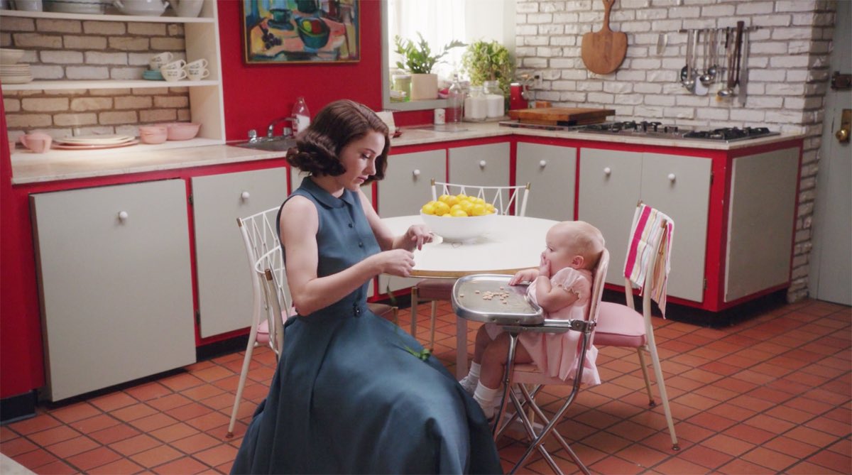Mrs. Maisel sitting in her 1950s-inspired kitchen alongside her baby who is sitting in a high chair.