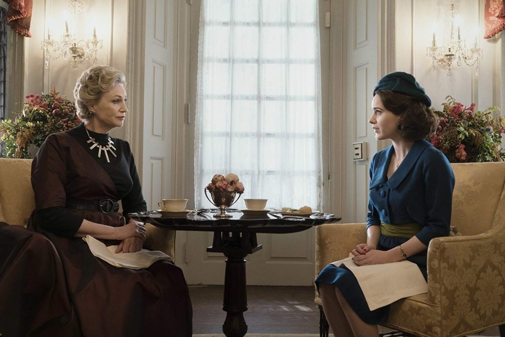 Mrs. Maisel sharing tea alongside a cast-member, both of whom are sitting in elegant patterned accent chairs.