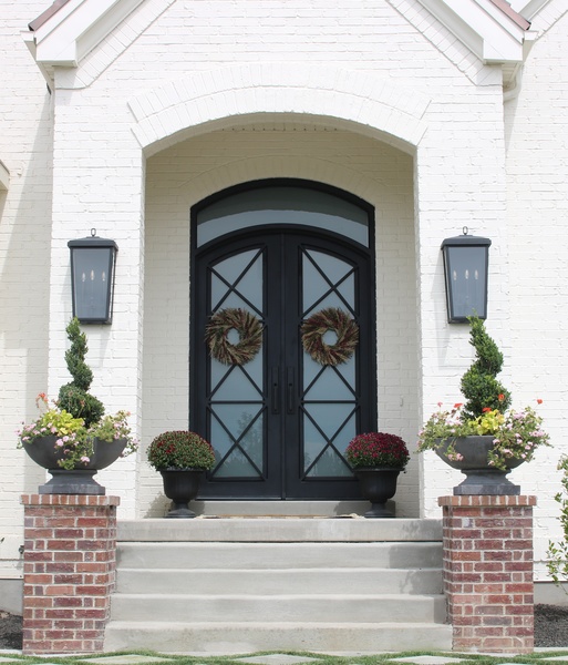 Capital Lighting's Donnelly Outdoor Lanterns flank the front arched door of this white brick home. Playing off the flat black finish on the steel doors, the lanterns feature a simple, traditional look that adds stately charm.