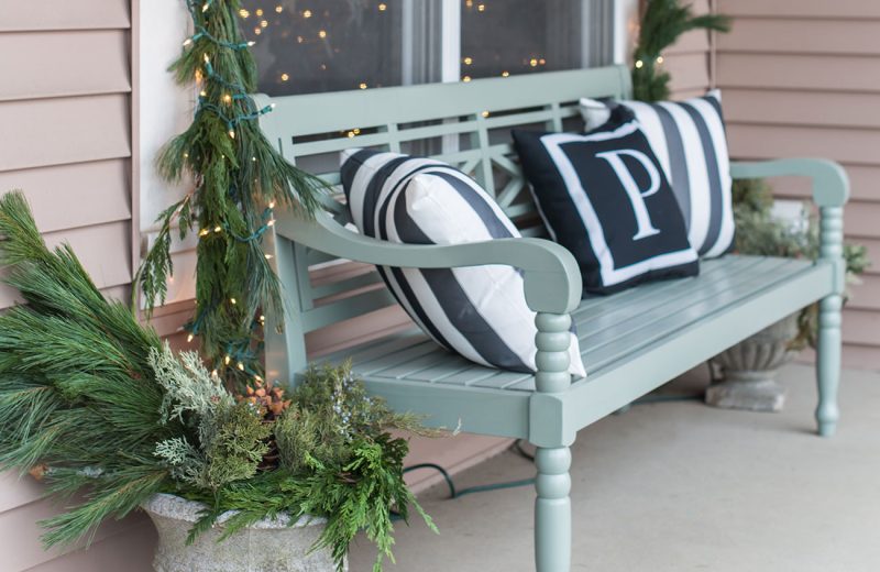 Fresh greenery Christmas decor with a garland around a window and a blue bench on the porch