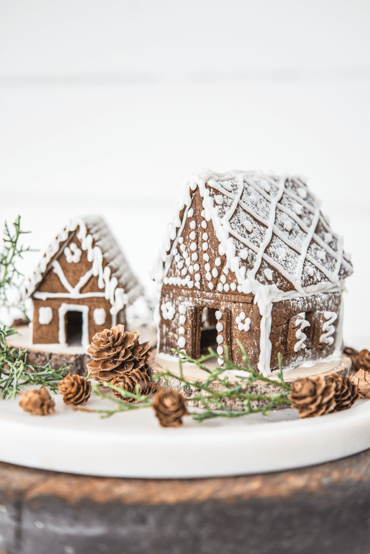 Two small gingerbread houses with white frosting details surrounded by pinecones and greenery. 