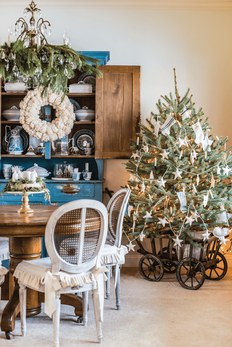 Fresh greenery hangs from the chandelier in Miss Mustard Seed's dining room. The Blue painted hutch features a yarn pom-pom wreath and the petite Christmas tree is placed nearby in an antique wooden wagon. 