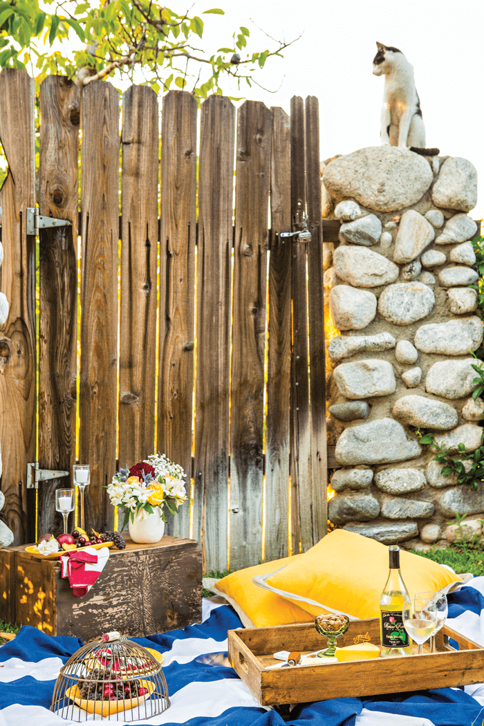 A cat sitting on a rock wall adjoined to a rustic wooden fence. A sprawling picnic set up on the ground below. 