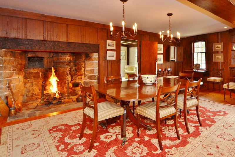 A dining room with a large rug and a large open fireplace lined with stone.