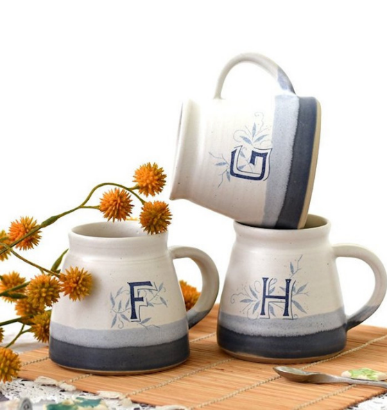 Stack of three mugs with blue detailing and monogram letter F, G and H on a table with yellow flowers