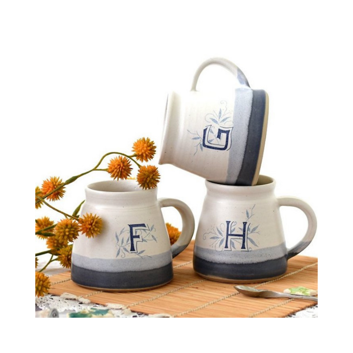 Stack of three mugs with blue detailing and monogram letter F, G and H on a table with yellow flowers