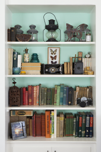 A bookshelf with the back painted light blue. Old books and other vintage accessories are displayed on it.