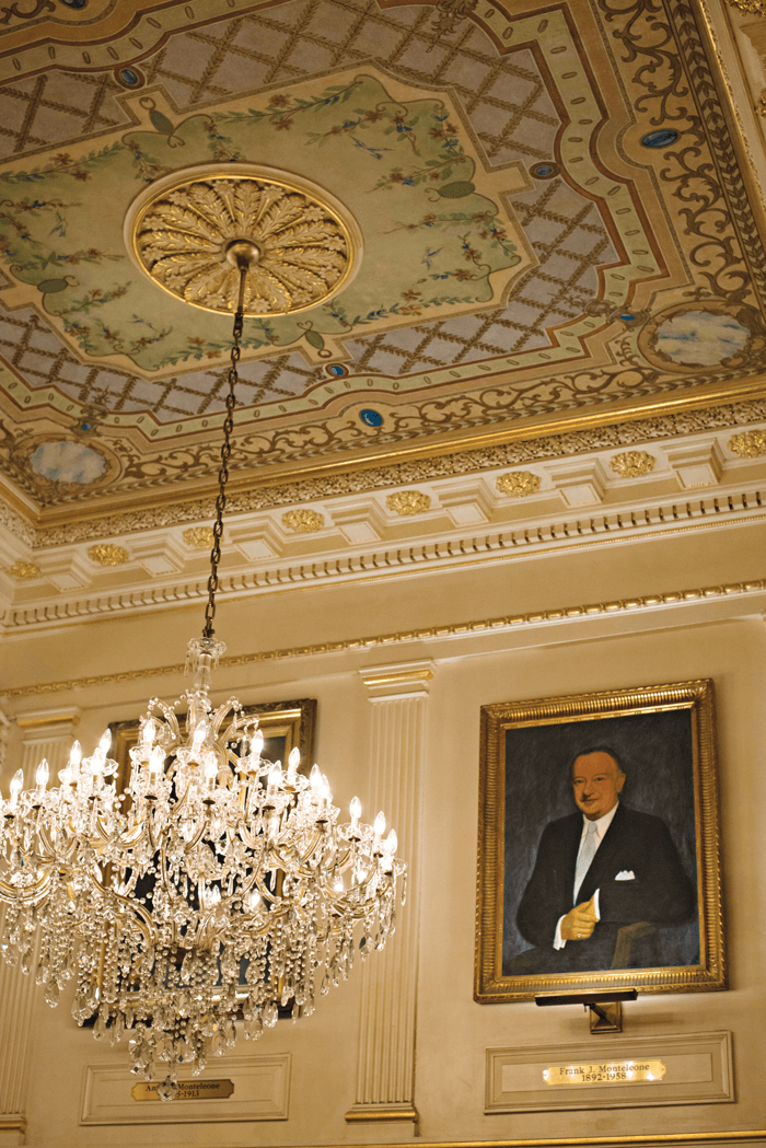 Crystal chandelier hanging from an ornate ceiling with a photograph of the hotel's founder, Antonio Montelone on the wall. 