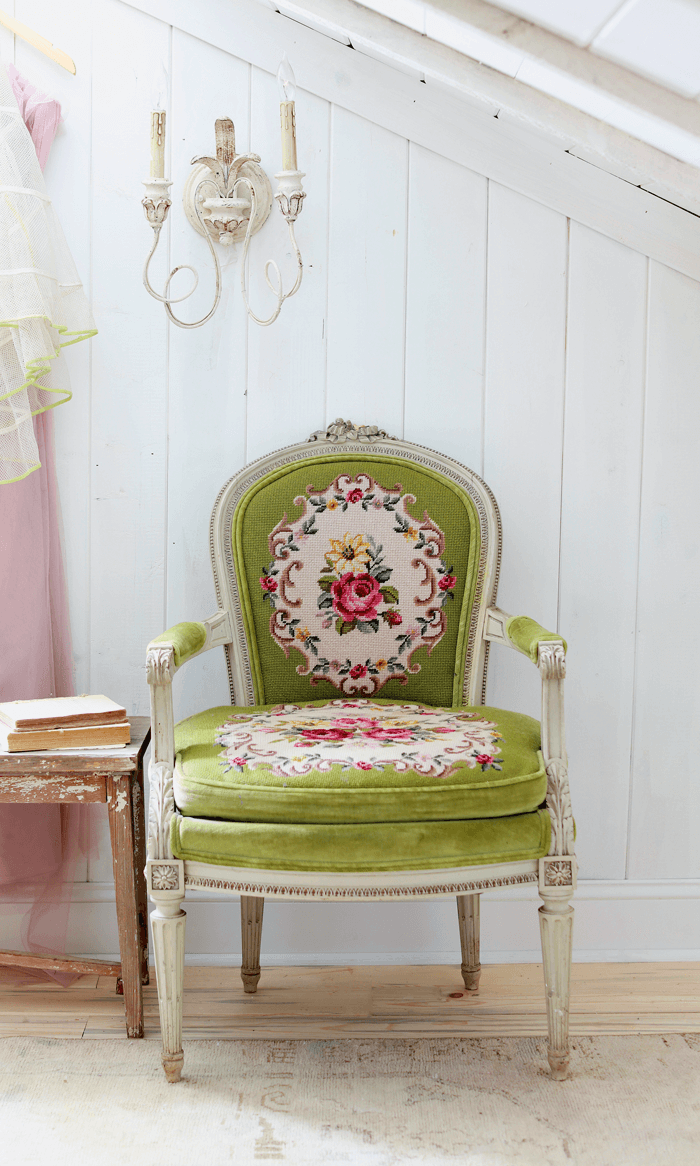 White vertical shiplap walls, stripped floors, ornate green needlepoint covered chair and a rustic wall sconce. 