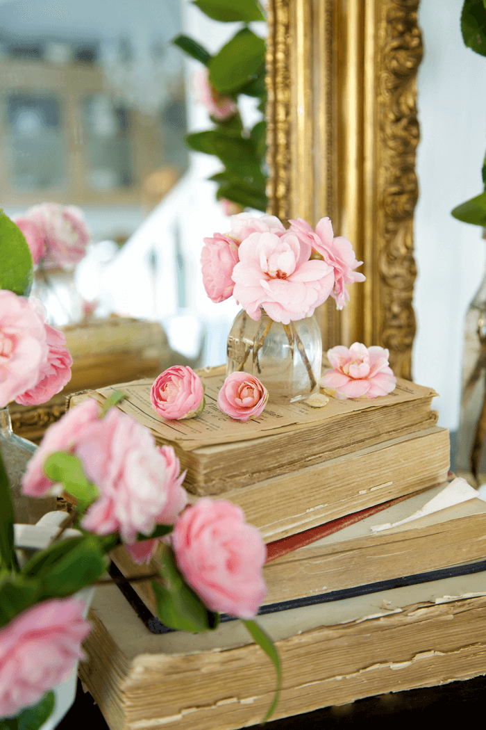 Stack of vintage books topped with pink peonies reflected in ornate gold-trimmed mirror. 