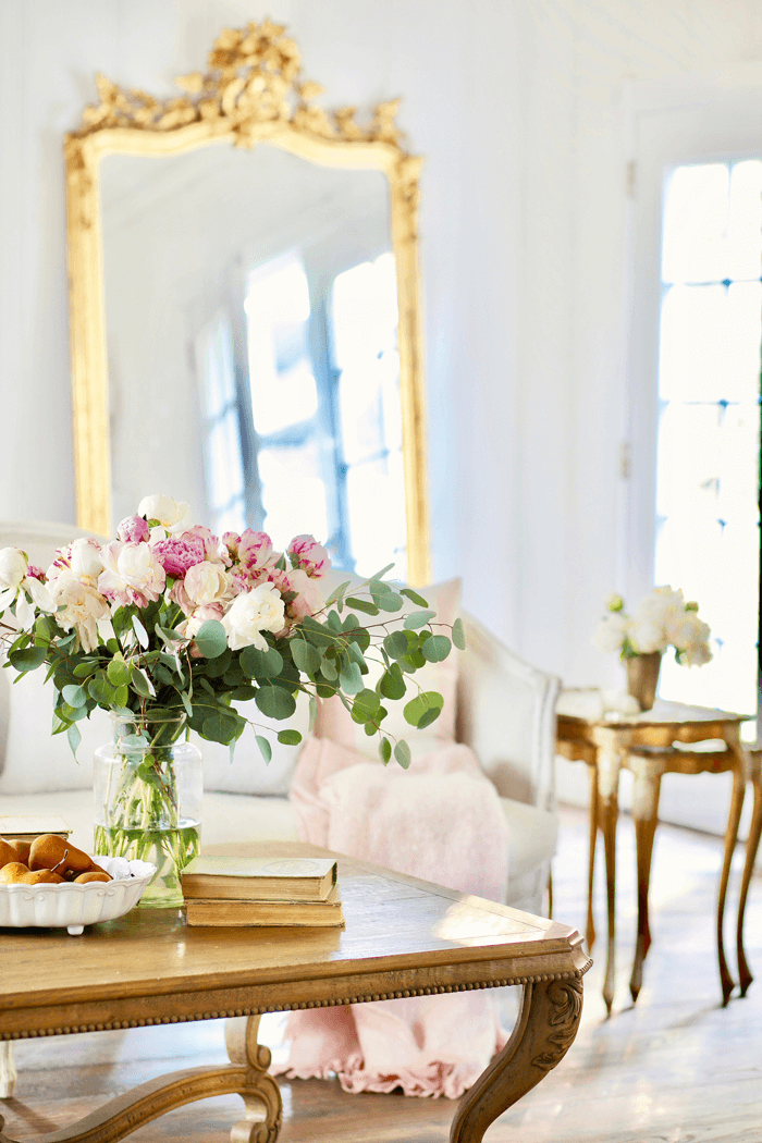 Wooden coffee table topped with vintage books and fresh flowers. White sofa and ornate floor to ceiling mirror in the background.