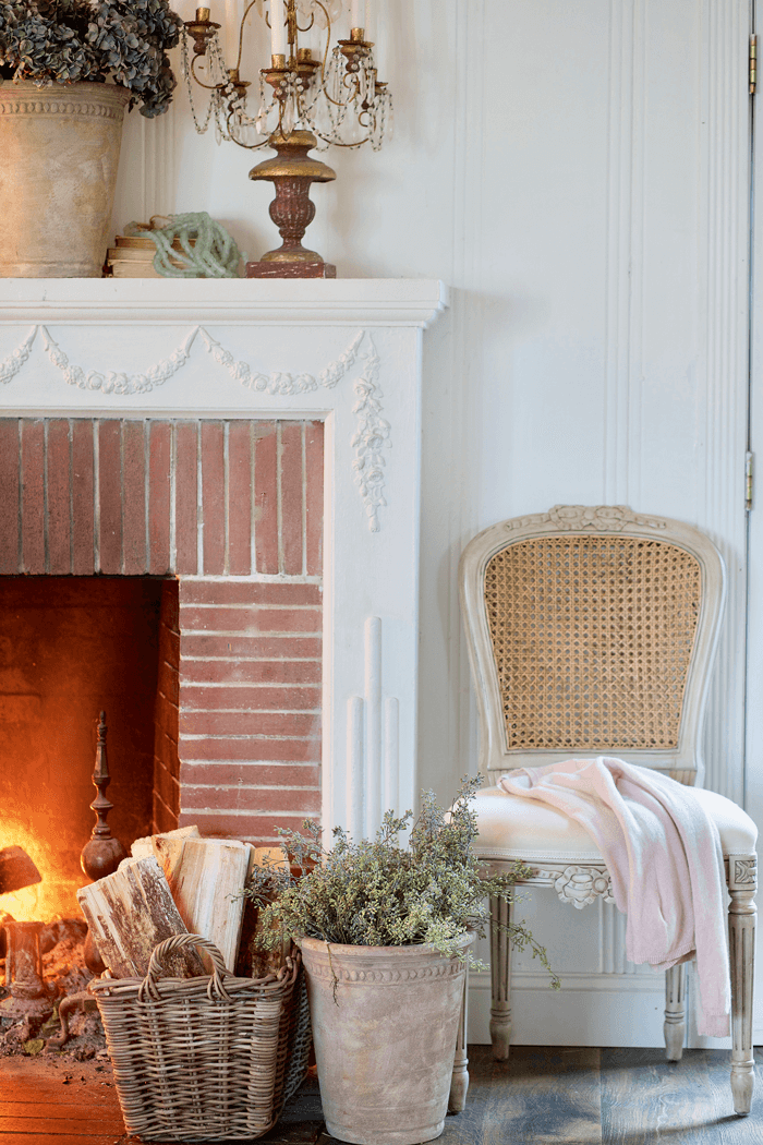 Fire lit in a brick fireplace. Fresh firewood in a basket in front of the fire situated next to a clay pot filled with greenery and a restored chair. Bronze candleabra with white candles on the mantle over the fireplace.