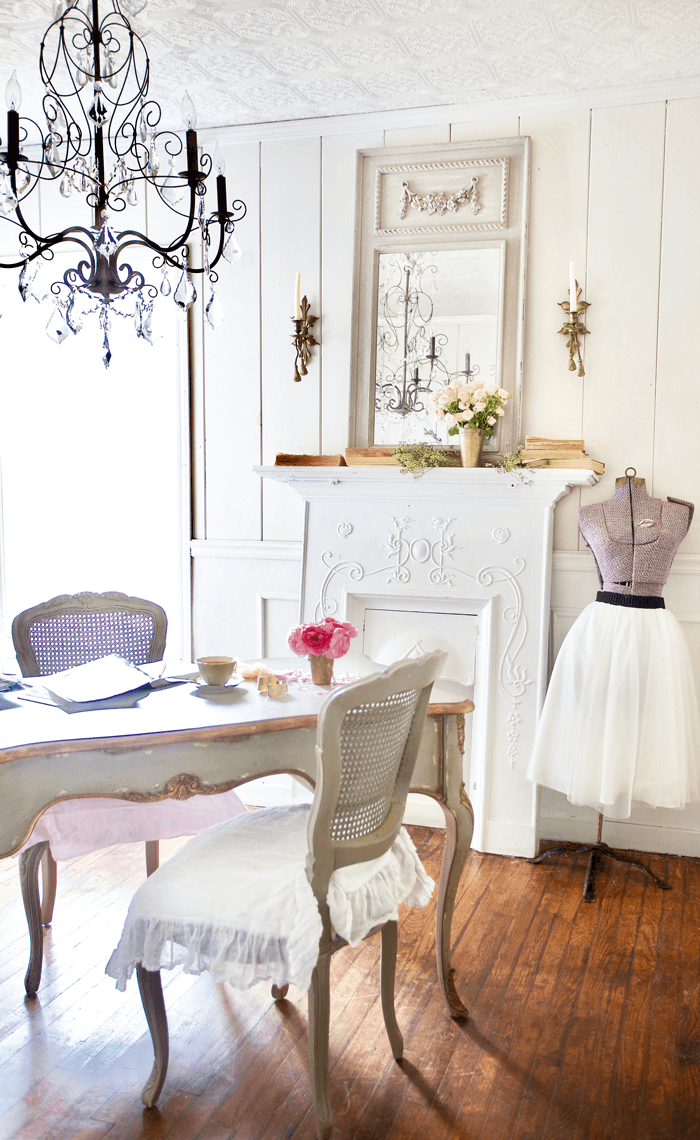 Romantic, rustic office with distressed light, bright desk and two chairs, sewing form and faux fireplace mantle. Hanging rod iron chandelier and bronze wall sconces on either side of a vintage mirror.