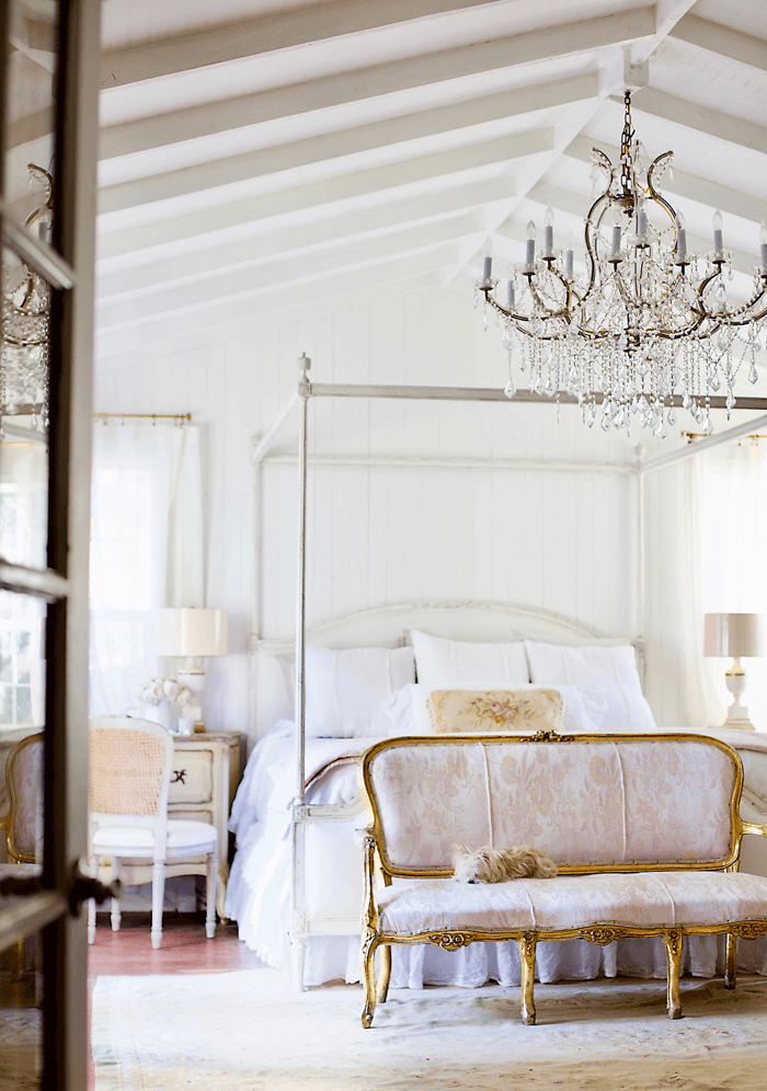 White four-poster bed sitting under a vaulted, exposed beam ceiling. Crystal chandelier hanging over the bed and gold rimmed bench at the end of the bed with a small, white dog curled up and asleep on it. 