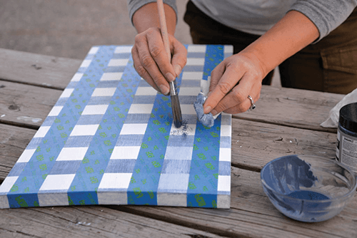 Hands painting into a gingham pattern with freshly placed tape in the opposite direction. 