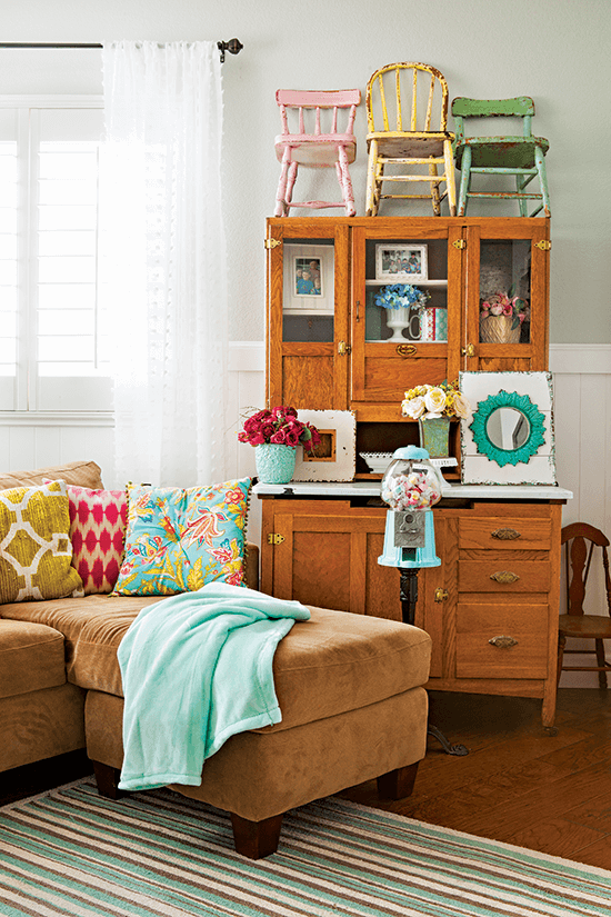 Tan microsuede sectional, brown wooden antique secretary desk covered with vibrantly colored wooden child chairs and featuring an heirloom bubblegum machine. 