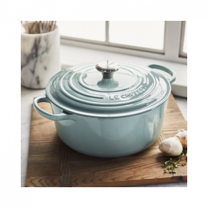 Soft blue colored Dutch oven with lid, laid out on wood cutting board next to fresh garlic and herbs.