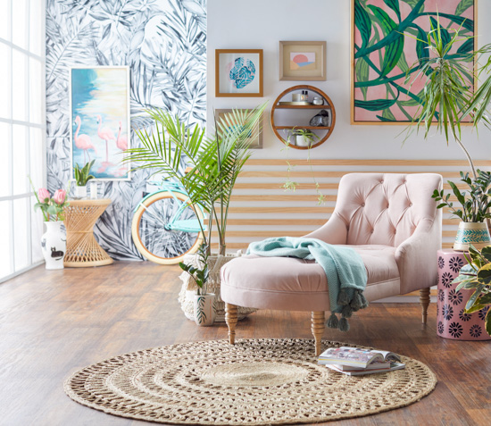 Jute round rug with a blush chaise lounge. Colorful and funky background walls with wallpaper as well as large art pieces, house plant and ceramics decorate the floor around the seating area.