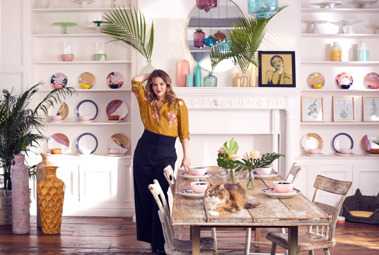 Drew Barrymore standing in a dining room setting. She's next to a rustic looking table with a tabby cat laying on top of it and her new collection is used as decor in the entire scene.