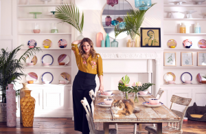 Drew Barrymore standing in a dining room setting. She's next to a rustic looking table with a tabby cat laying on top of it and her new collection is used as decor in the entire scene.