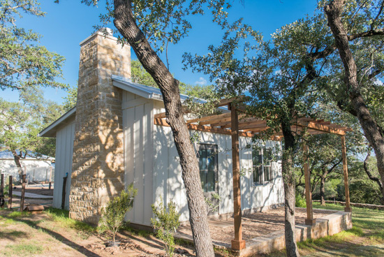 Cottage with wooden pergola and stone fireplace surrounded by trees.