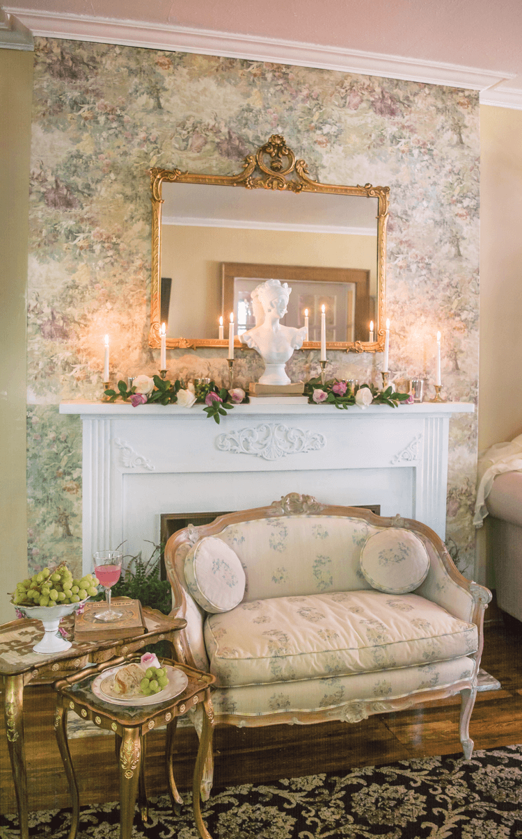 accent wall wallpapered in floral print with a white mantle and gold accented mirror with lovely seating in front. 