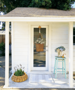 The view of the front porch and door to the she shed, white shiplap and welcoming with a striped mat and fresh flowers.