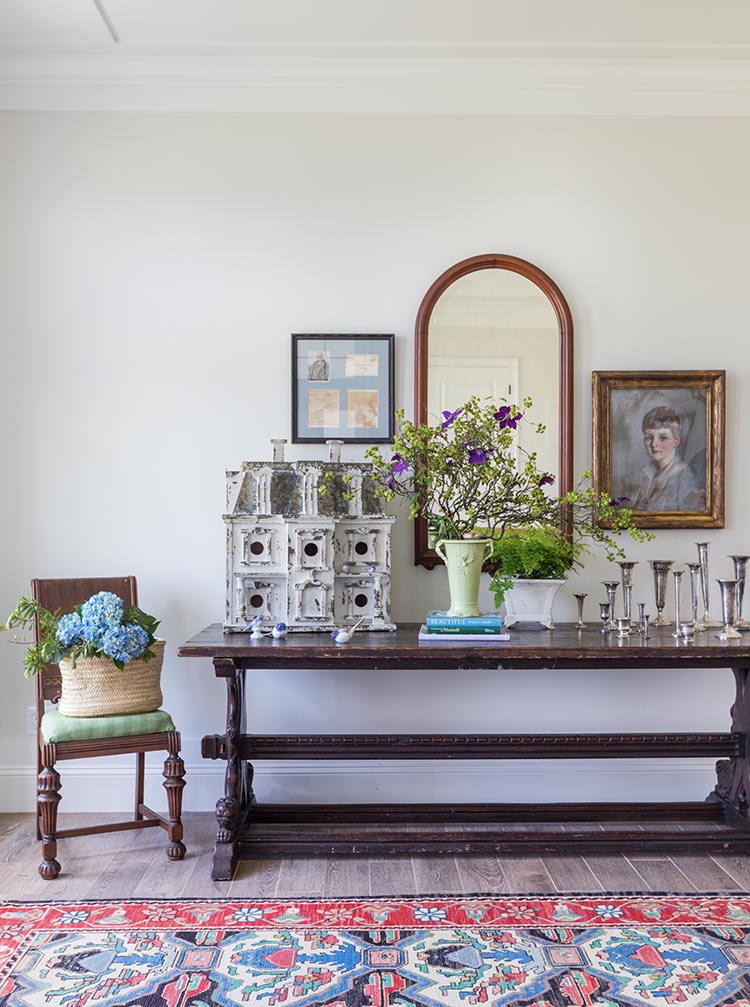 A vintage refectory table with a birdhouse and silver vases on it. A mirror and portrait hang above.