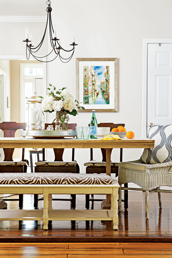 A view of the eclectic dining table set with a wicker chair, three dark wooden chairs and a light colored zebra printed upholstered bench for seating. 