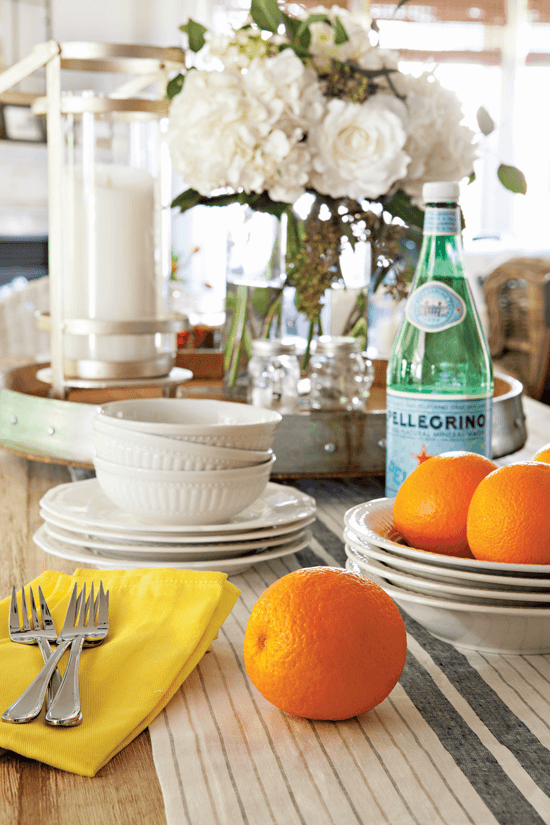 The dining table laid out with crisp and clean white dishes. Forks, bowls and sparkling water are set out with some fresh oranges on display for color and coziness. 