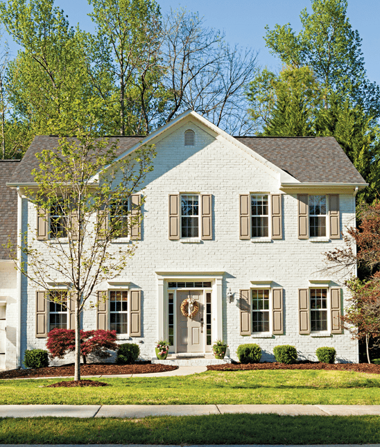 Front fascade of a home. Was old red brick, now painted light cream and the black shades also painted a light color with mature trees growing the back yard and showing over the top of the second story.