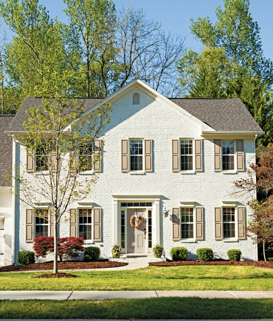 Front fascade of a home. Was old red brick, now painted light cream and the black shades also painted a light color with mature trees growing the back yard and showing over the top of the second story. 