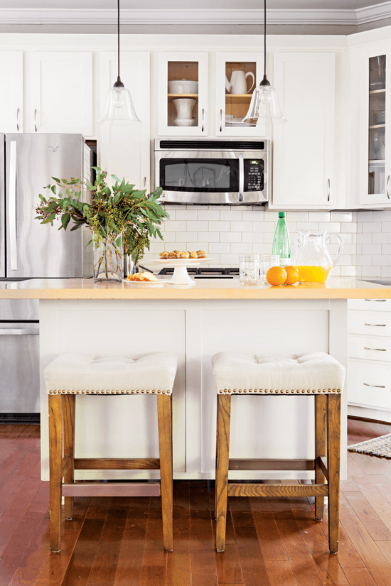 Looking over the upholstered bar stools and kitchen island at the freshly painted light and bright kitchen cabinets and appliances. 