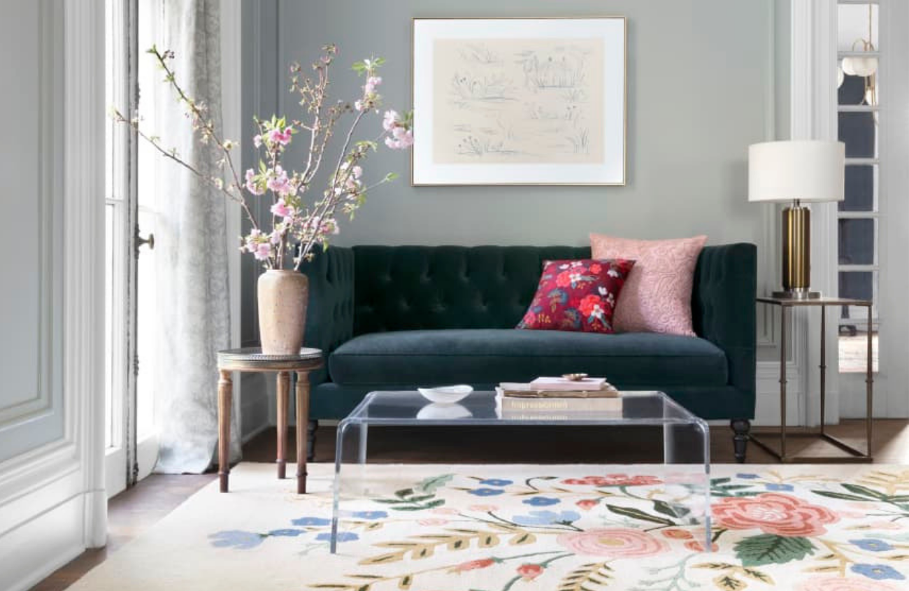 Living room scene with clear coffee table. pink flowers in a vase, a gem toned velvet couch and featuring Rifle Paper Co. pillow and rug with floral design.